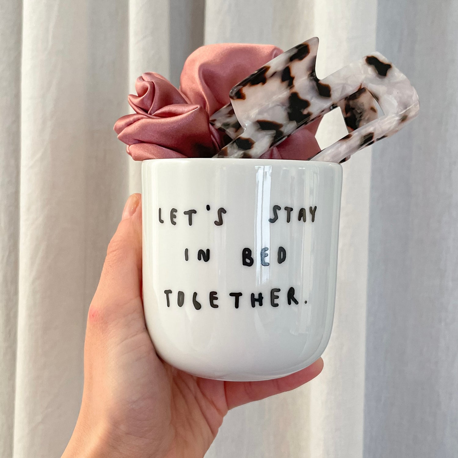 Keramikbecher mit der Aufschrift "Lets Stay in bed together". In diesem befindet sich eine Haarklammer im Leo-Muster und ein pinker Haargummi aus Satin